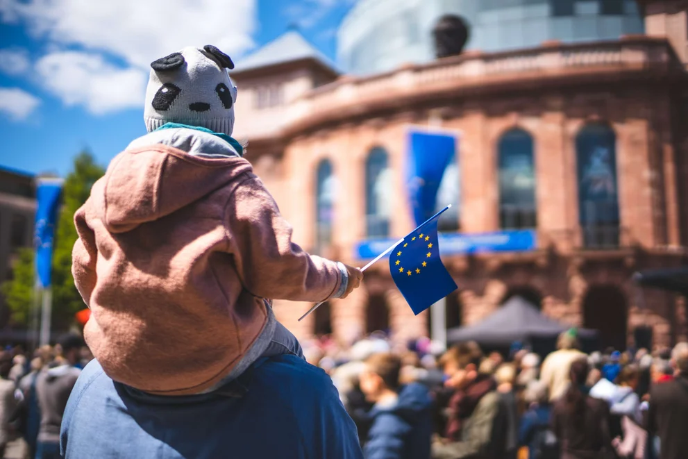 Ein Kind hält eine kleine Europa-Fahne in der Hand und sitzt auf der Schulter einer erwachsenen Person. 