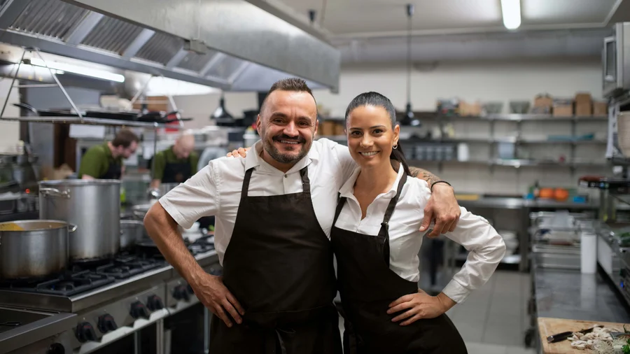 Zwei Betreiber:innen einer Gaststätte stehen in der Küche und lächeln