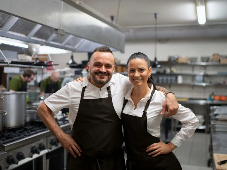 Zwei Betreiber:innen einer Gaststätte stehen in der Küche und lächeln