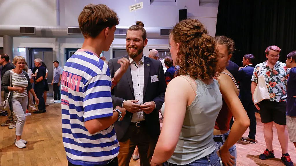 Unser Abgeordneter Yannick Veits im Gespräch mit Bürger:innen und steht dabei in einem großen Saal.