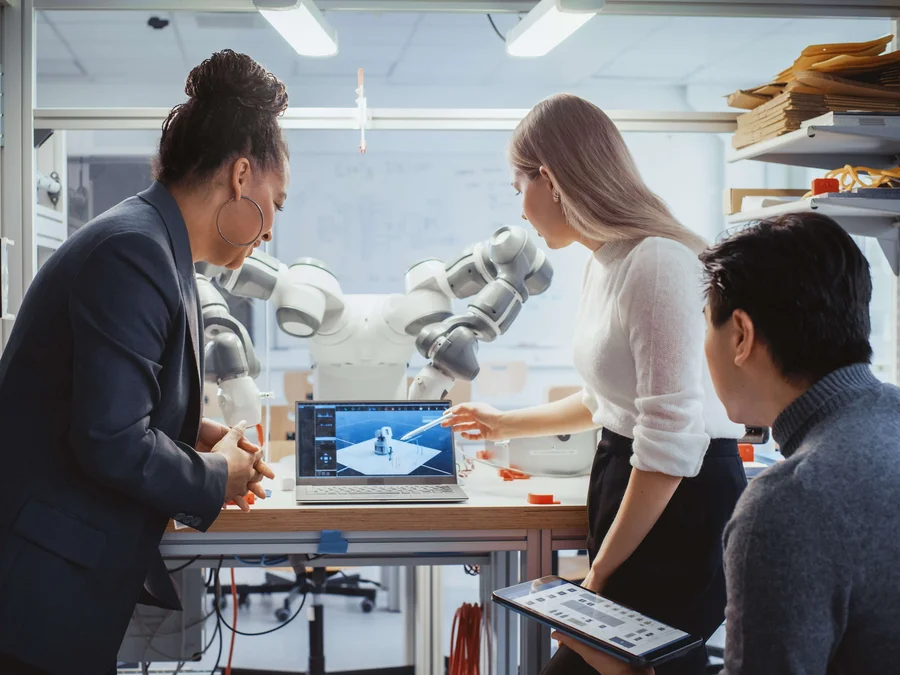 Diverses Team von Studierenden arbeiten an der Foschung zur Robotik in einem technischen Labor. Im Hintergrund ist der Roboter zu erkennen. 