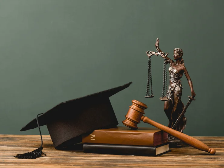 Ein Abschlusshut und ein Gerichthammer liegen auf einem Gesetzbuch auf einem Tisch. Im Hintergrund steht eine Justicia-Figur.