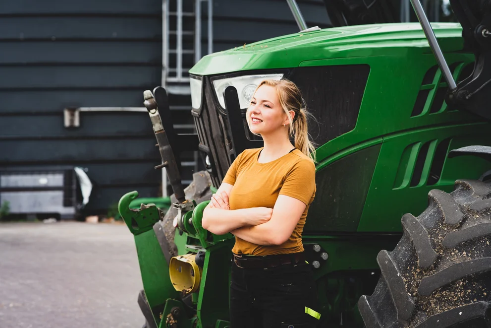 Glücklich wirkende, weiblich gesehen Person steht vor einem grünen Traktor. 