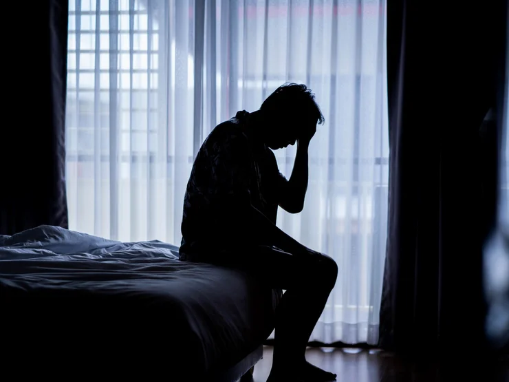 Mann sitzt erschöpft auf einem Bett in einem abgedunkelten Zimmer.