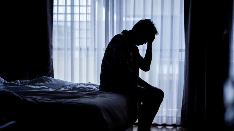 Mann sitzt erschöpft auf einem Bett in einem abgedunkelten Zimmer.