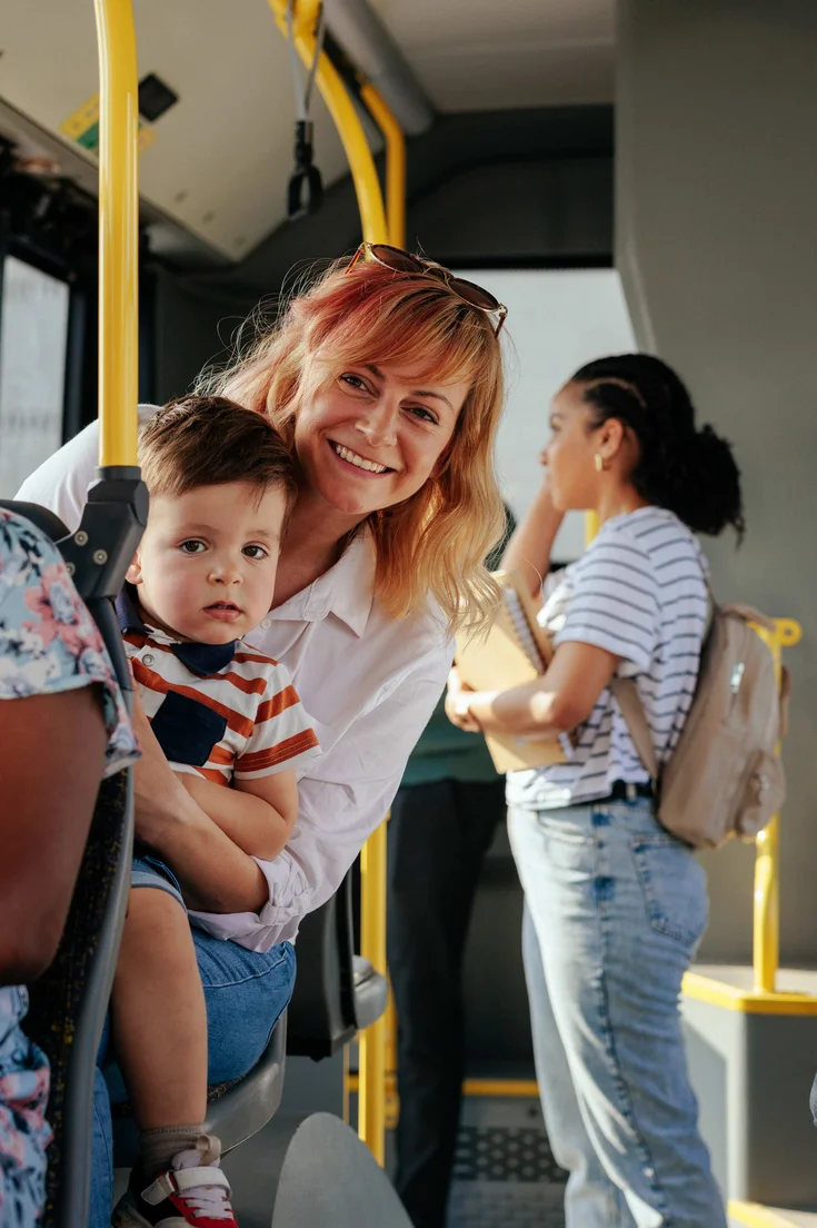Eine Person hat ein Kleinkind auf dem Schoß während einer Busfahrt und lächelt dabei.