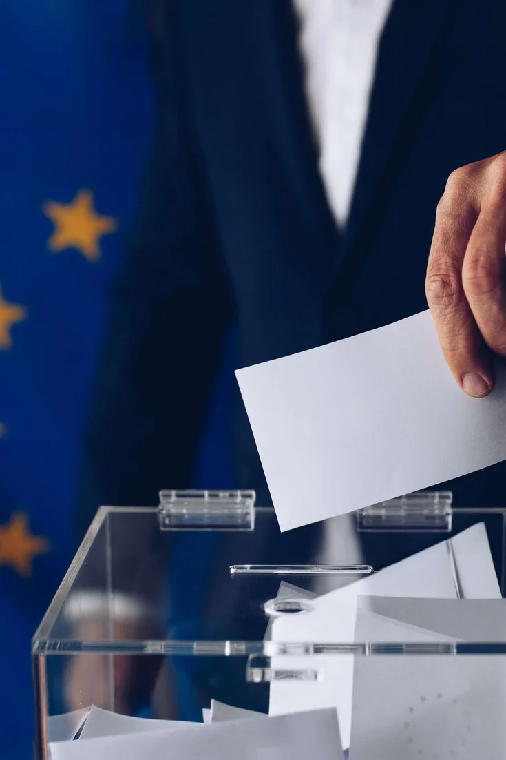Eine Person in einem blauen Anzug steckt einen Wahlzettel in eine Wahlurne. Im Hintergrund ist eine Europa-Fahne zu sehen.